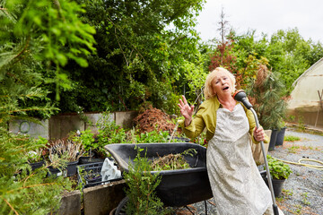 Elderly woman as a gardener sings into a hose for fun
