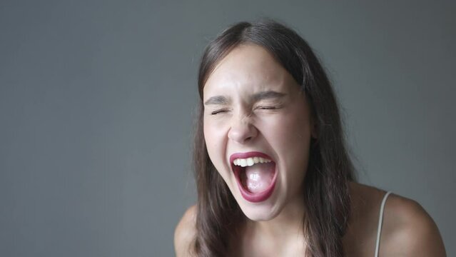 portrait of screaming loud young brunette female with red lips wide opened mouth, closed eyes. dark gray background, facial expression mad, yelling, shouting emotions. caucasian woman face close up