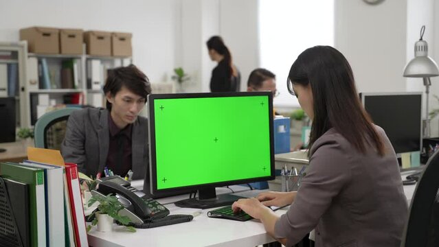 backside view of Japanese female worker wearing suit is using desktop computer with green screen while working with other coworkers as a team in the office
