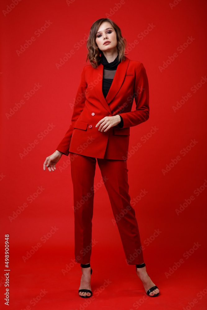 Wall mural High fashion photo of a beautiful elegant young woman in a pretty red suit, jacket, pants, trousers, black blouse posing on background. Slim figure, hairstyle, studio shot. Monochrome, total red