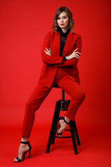 Naklejka premium High fashion photo of a beautiful elegant young woman in a pretty red suit, jacket, pants, trousers, black blouse posing on background. Monochrome, total red. Model sits on chair