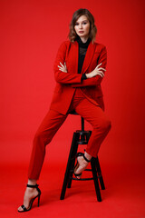 High fashion photo of a beautiful elegant young woman in a pretty red suit, jacket, pants, trousers, black blouse posing on background. Monochrome, total red. Model sits on chair