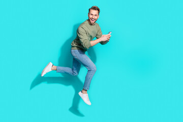 Fototapeta na wymiar Full length photo of cheerful influencer man jump hold telephone run wear pullover isolated blue color background
