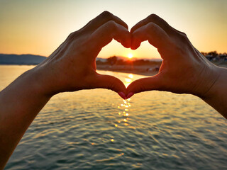 Senior woman hands in heart shape love sign around sun during sunset on the beach.