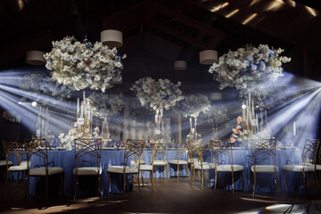 Round dining tables, covered in d blue cloth, served with porcelain and blue and purple glasses and richly decorated with flowers, flowers under the tables. Bokeh gold candles on