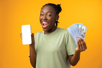 Young black woman holding white screen smartphone and money in yellow studio