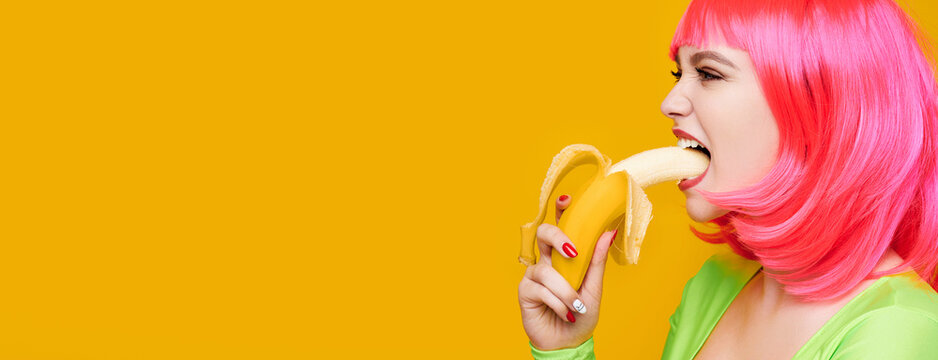 Sexy hipster woman in pink wig eat banana on bright yellow background