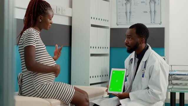 Doctor using greenscreen dispay on digital tablet at checkup visit with pregnant woman. Health specialist showing mockup template with isolated chroma ley and blank copyspace to expectant patient.