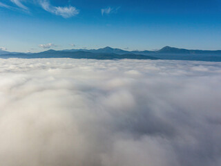 Obraz premium Flight over fog in Ukrainian Carpathians in summer. A thick layer of fog covers the mountains with a solid carpet. Mountains on the horizon. Aerial drone view.
