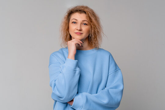focused middle age woman with curly blonde hair looking for answers on her questions, touching chin, standing in trendy blue sweatshirt over light grey background