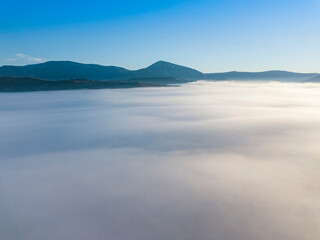 Obraz premium Flight over fog in Ukrainian Carpathians in summer. Mountains on the horizon. A thick layer of fog covers the mountains with a continuous carpet. Aerial drone view.