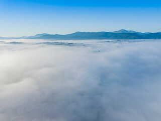 Obraz premium Flight over fog in Ukrainian Carpathians in summer. Mountains on the horizon. A thick layer of fog covers the mountains with a continuous carpet. Aerial drone view.