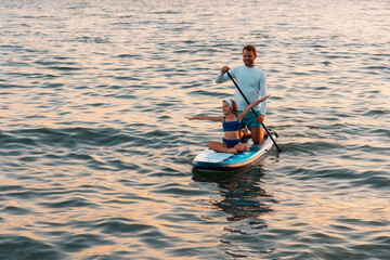 Father and daughter surfing on a sup board. Sea at the background. Summer vacations