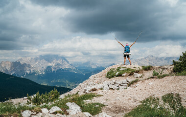 Woman trekker with backpack rose arms up with trekking poles enjoying picturesque Dolomite Alps...