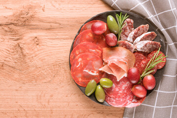 Top view on charcuterie plate with different types of sausages - salami, bresaola, proscuitto served with olives and grape. Black plate with traditional italian antipasti on wooden table 
