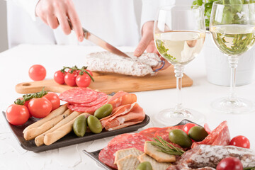 Obraz na płótnie Canvas Woman serving traditional Italian antipasti on white table. Charcuterie plate with different types of sausages - salami, bresaola, proscuitto served with olives, baby tomatoes