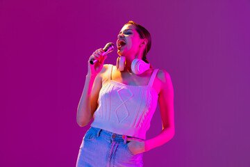 Portrait of young beautiful happy girl singing with microphone isolated on purple background in neon light, filter. Concept of emotions, music, facial expressions
