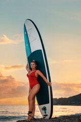 Plus sized sexy tanned woman in red bikini poses with sup board at beach. Sunset sky on the background. Vertical orientation. The concept of surfing and summer vacation
