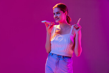 Young beautiful happy girl recording voice message on phone isolated on purple background in neon filter. Concept of emotions, digital technologies, fashion