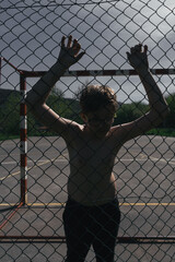 BOY WITH GLASSES AND NO SHIRT IS RESTING LEANING ON THE FENCE OF A SOCCER FIELD. BEHIND IS A RED AND WHITE GOAL.