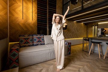 Young woman stretching up, waking from a sleep while standing at modern living room at home. Concept of home comfort and good morning. Caucasian woman wearing sleepwear
