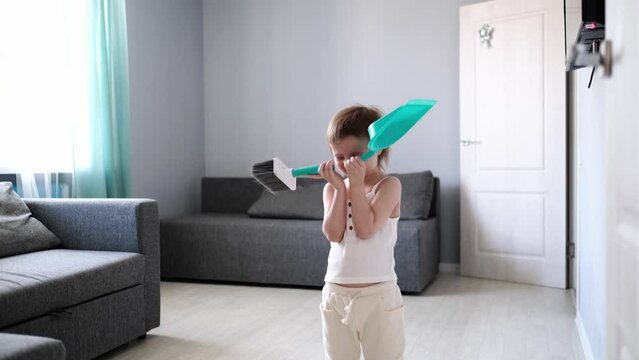 Toddler girl cheerfully sweeps the floor with a brush and dustpan. The child dances and cleans the room, lifestyle in a real interior