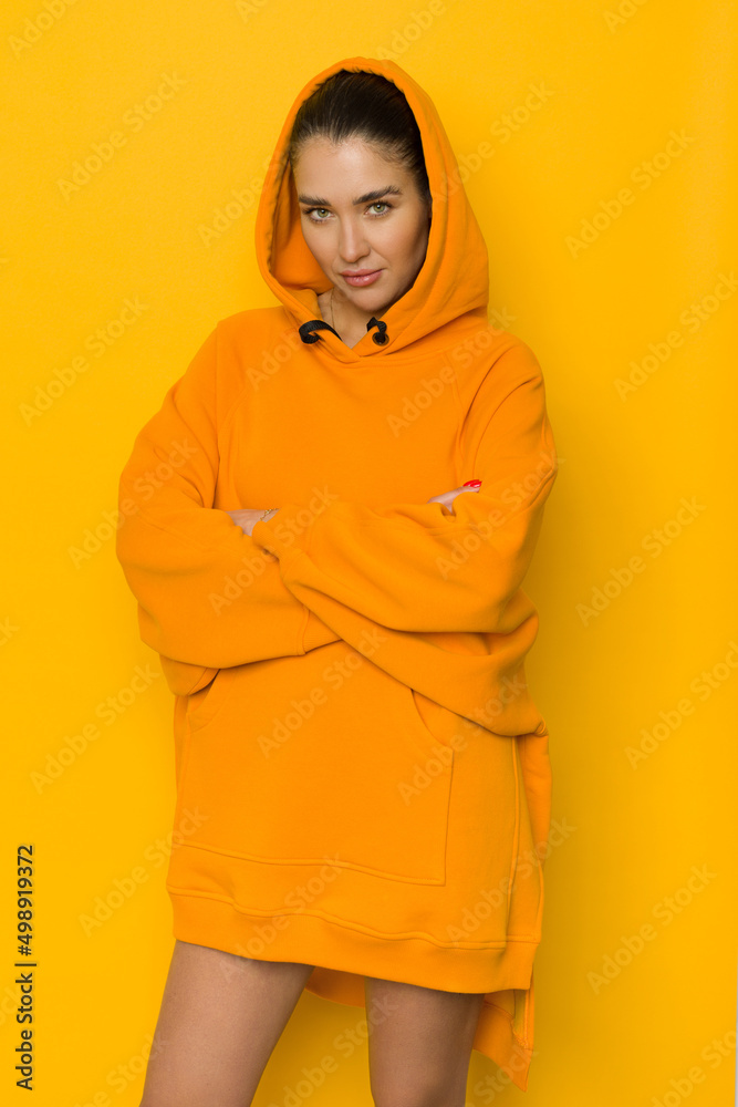 Wall mural confident young woman is posing with arms crossed in orange oversized hooded sweatshirt.