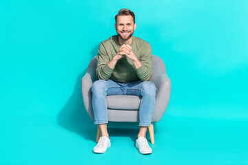 Fototapeta na wymiar Photo of positive cheerful man sit armchair hold hands beaming shiny smile wear pullover isolated blue color background