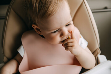 10 months old baby sitting in high chair, with a silicone bib, eating finger food.