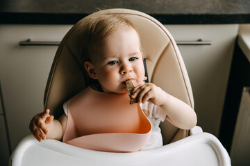 10 months old baby sitting in high chair, with a silicone bib, eating finger food.