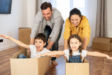So fun. Happy family relocated in new home, unpacking together, overjoyed middle-aged parents are pushing two kids sitting and riding in cardboard boxes. New property and relocation - Powered by Adobe