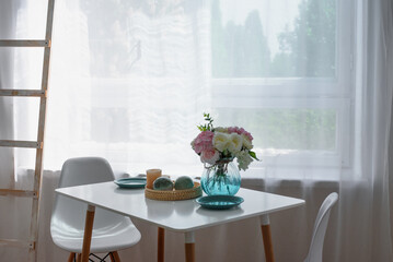 White kitchen table against the window. On the table are plates and a vase of flowers. Contrasting photo.