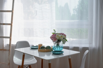 White kitchen table against the window. On the table are plates and a vase of flowers. Contrasting...