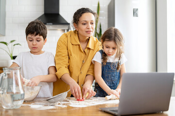 Middle-aged mother and her kids cooking together, kneads the dough, using rolling pin. Mom and kids spends leisure at home, baking cookies, looking tutorial at the laptop, enjoying weekend activity