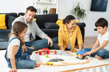 Mom, dad and children sitting on floor in cozy living room and playing. Lovely family of four spend weekend together at home, parents and two kids playing toys, sibling building railway, have a fun
