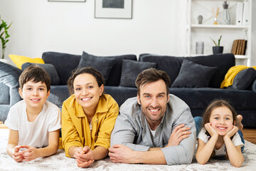 Portrait of cheerful multiracial family of four - mom, dad, son and toddler daughter laying at the floor in cozy living room, looking at camera and laughing, enjoying weekend at home