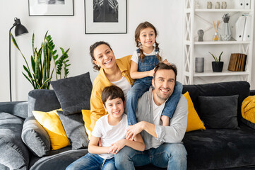 Family harmony. Happy multiracial family of four sitting on the sofa in cozy living room, looking at the camera and smile. Portrait of cheerful spouses and two kids