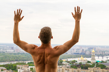 A muscled man is staying and training, keeping the hands up, body of muscleman, strapper, bodybuilder on a roof with amazing view on central part of city in Kyiv