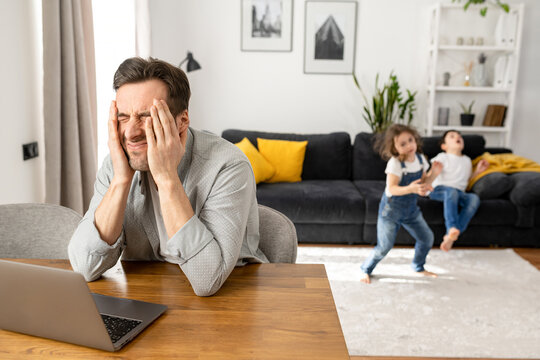 Annoyed father sitting with laptop with eyes closed, covering head with hands while his two kids are jumping and shouting around. Irritated freelancer parent is babysitting and working