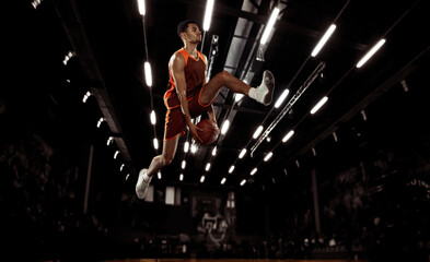 Fototapeta na wymiar In action. Young basketball player jumping with ball in flashlights over dark gym background. Concept of sport, energy and dynamic, healthy lifestyle. Arena's drawned