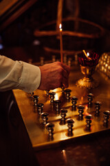 Unrecognizable man wearing white shirt, putting burning candle on golden square stand with candlesticks in orthodox church. Faith in God, religion, ceremony, praying, holiday, commemoration concept.