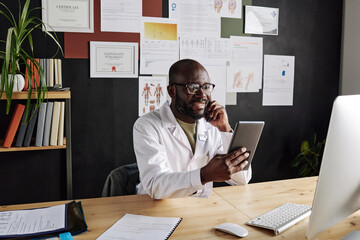 African male doctor sitting at workplace holding tablet pc and smiling while talking on mobile phone