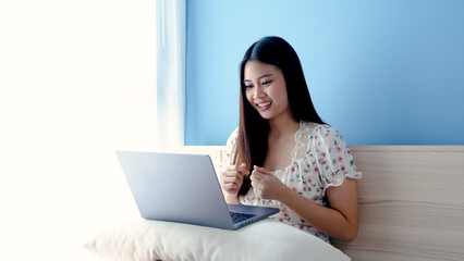 Naklejka premium Beautiful Asian girl, wearing a floral shirt, is ordering online on a laptop computer, happily, holding a credit card in her hand, picking up to pay with a credit card,online shopping.