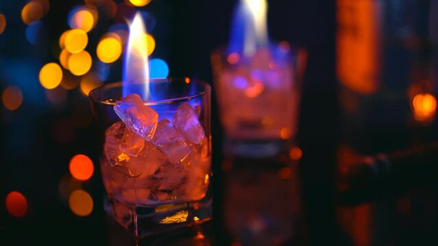 Burning ice in two glasses with strong alcohol on the bar counter. Colored lights of the bar illumination, a cigar and a bottle of white strong drink. Orange and blue flames in glasses with ice.