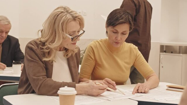 Waist up slowmo of two Caucasian mature women discussing Math problem while looking at paper practice test, sitting together at desk in classroom