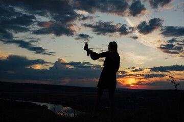 Dark silhouette of a woman takes a selfie photo on a smartphone while standing on the hill at sunset, evening.