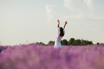 Young woman in a white dress standing with hands up in the middle of a lavender field in the sunny...