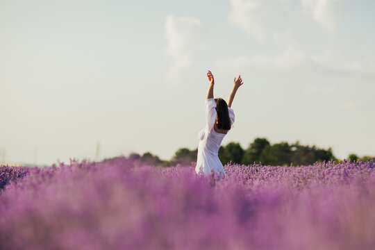 Young woman in a white dress standing with hands up in the middle of a lavender field in the sunny summer day.
