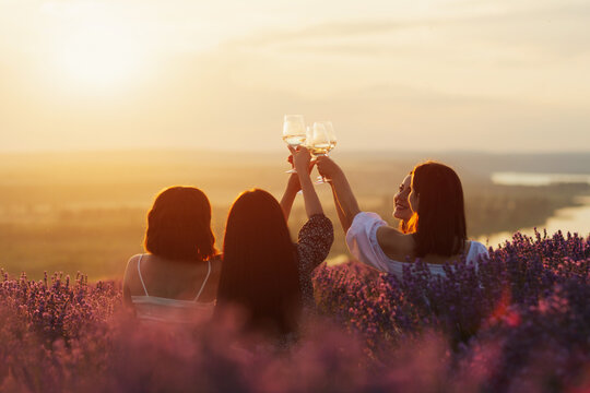 Friends having picnic in the lavender field. Group of young women sitting on lavender field at sunset on summer day. Girlfriends  drinking wine on outdoor party.