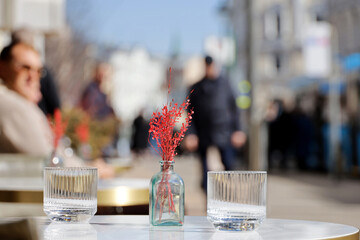 Defocused view to people in street cafe, decorative flowers and glasses on table outdoor. Romantic dinner in spring or summer city, elegant setting